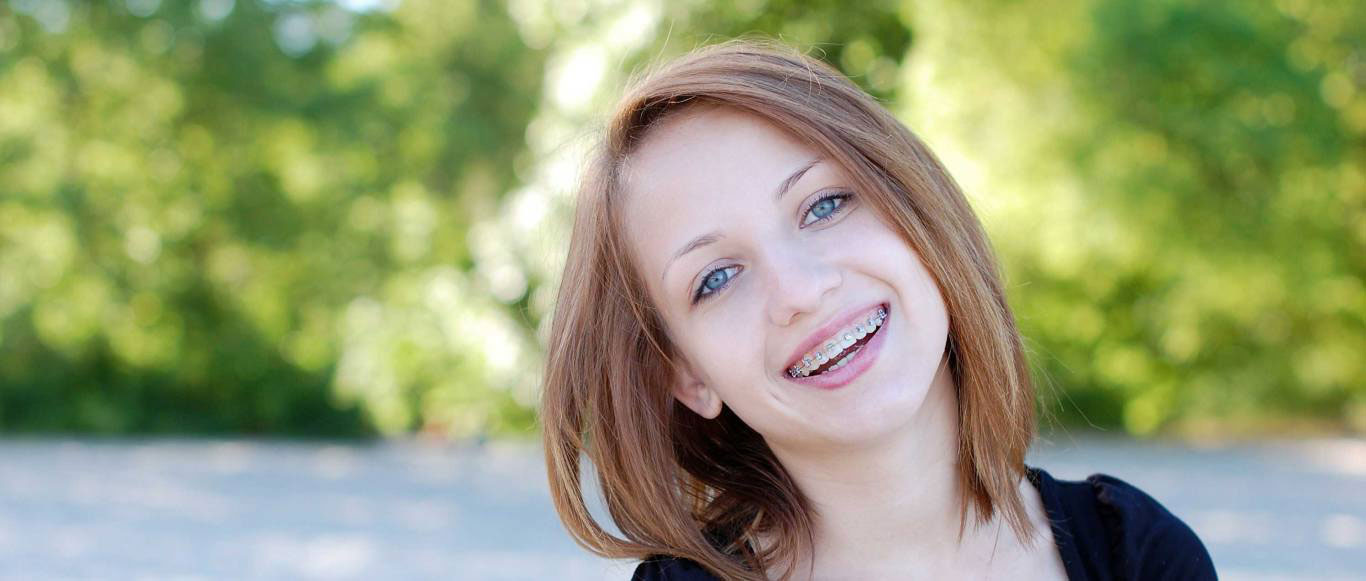 The image shows a young woman with short hair smiling at the camera, set against a blurred outdoor background.