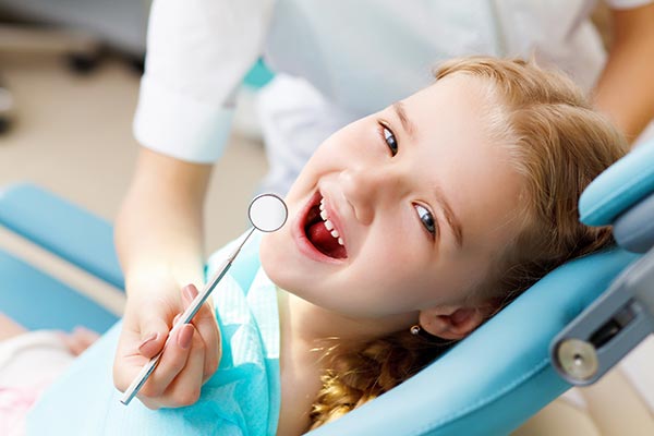 A young child sitting in a dental chair with a dental mirror in hand, smiling at the camera.