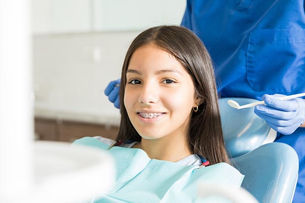 A young woman sitting in a dental chair with a smiling expression, receiving dental care from a dental professional wearing protective gloves and a blue gown.