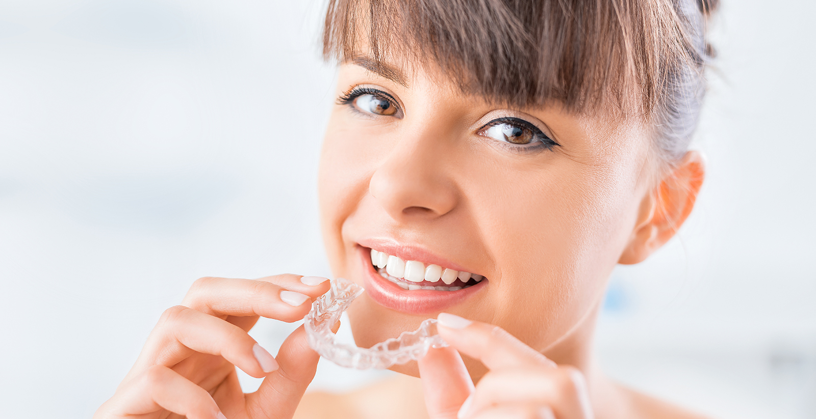 The image shows a smiling woman with an open mouth, holding a toothbrush near her teeth, which are covered with toothpaste foam, against a blurred background.