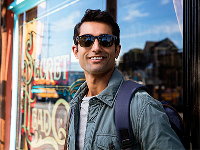 The image shows a man wearing sunglasses and a backpack standing outside a storefront with a sign that reads  Sweet Tooth.  He appears to be posing for the photo with a slight smile.