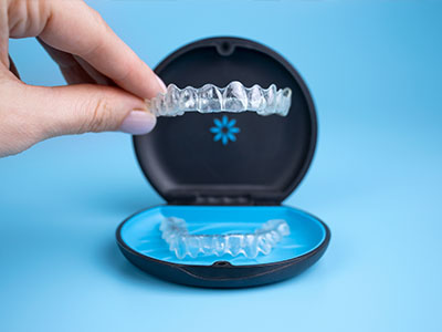 The image shows a person s hand holding a transparent plastic dental retainer with teeth-like ridges, placed inside an open blue case, which is commonly used for storing dental appliances.