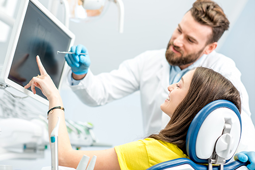 In the image, there are two individuals  a man standing next to a woman who is seated on a chair with a medical device attached to her head. They appear to be in a professional setting, possibly a dental or medical office, with various equipment and screens in the background. The man is wearing a white lab coat and has a stethoscope around his neck, suggesting he may be a healthcare professional.